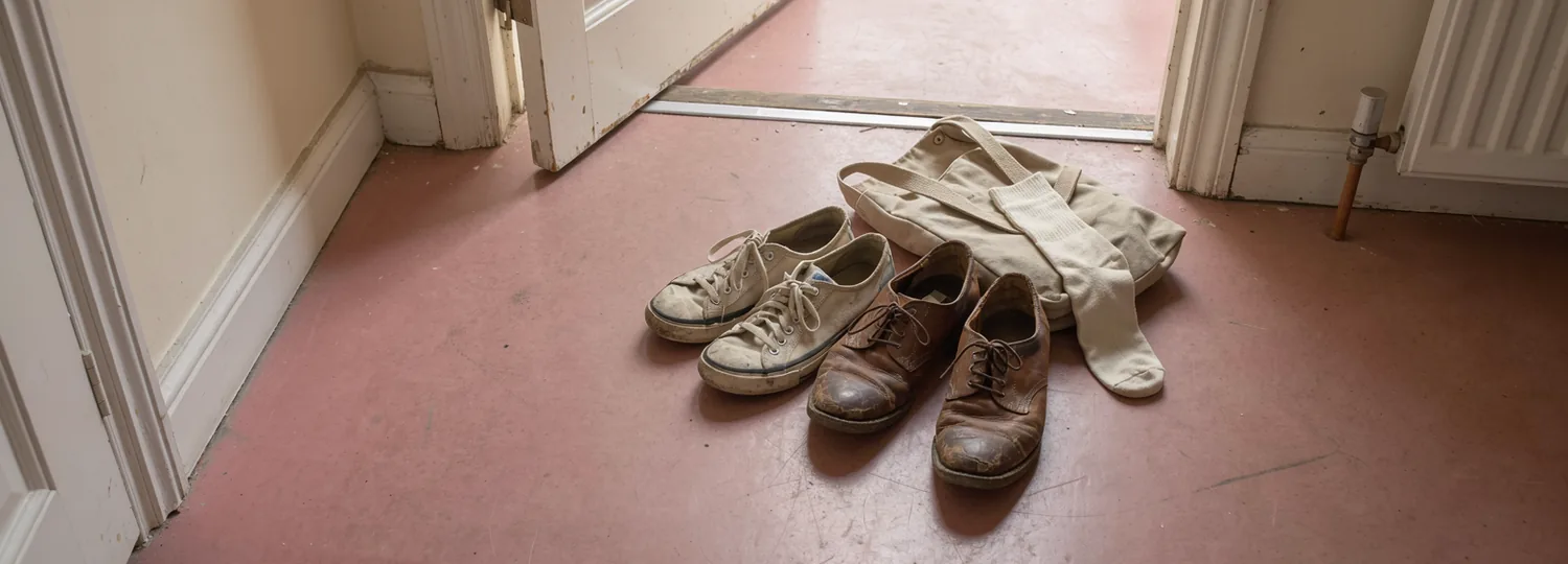 Warm daylight reaches an open doorway, two worn pairs of shoes and a canvas bag on an ordinary pink hallway floor.